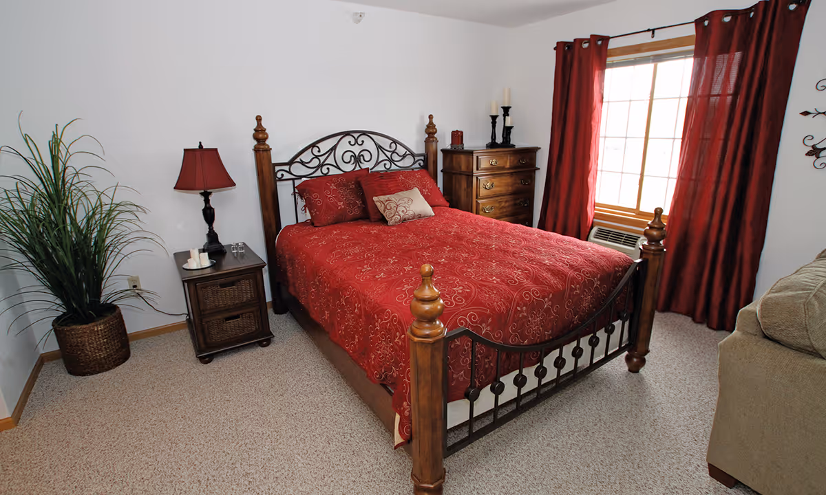 Well-lit bedroom with a wooden and metal-framed bed dressed in red bedding, a nightstand with lamp, dresser, potted plant, and a window with red curtains.