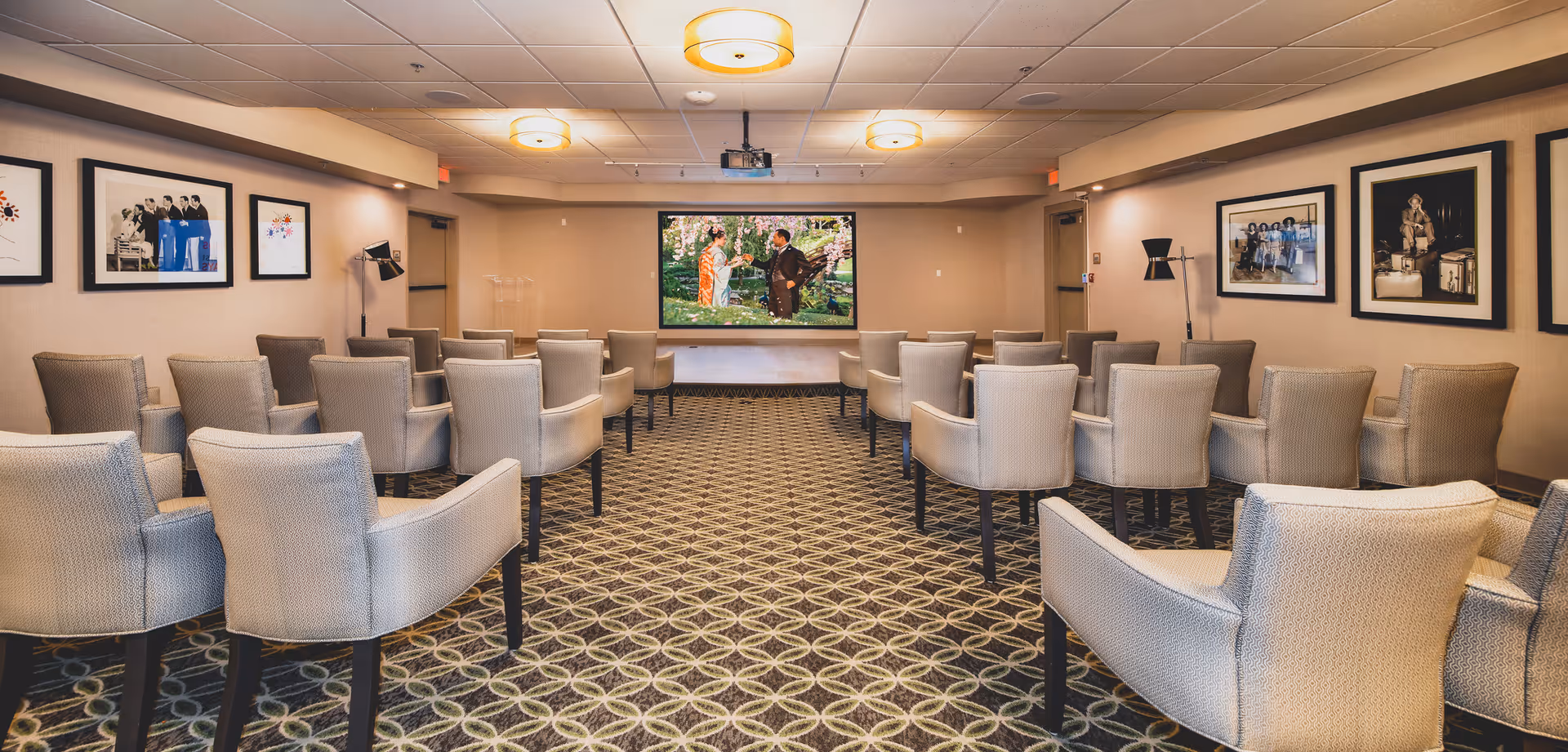 A small theater or presentation room with rows of beige upholstered chairs facing a large screen displaying a scene with two people outdoors. The room has patterned carpet, framed pictures on the walls, ceiling lights, and a projector mounted on the ceiling.