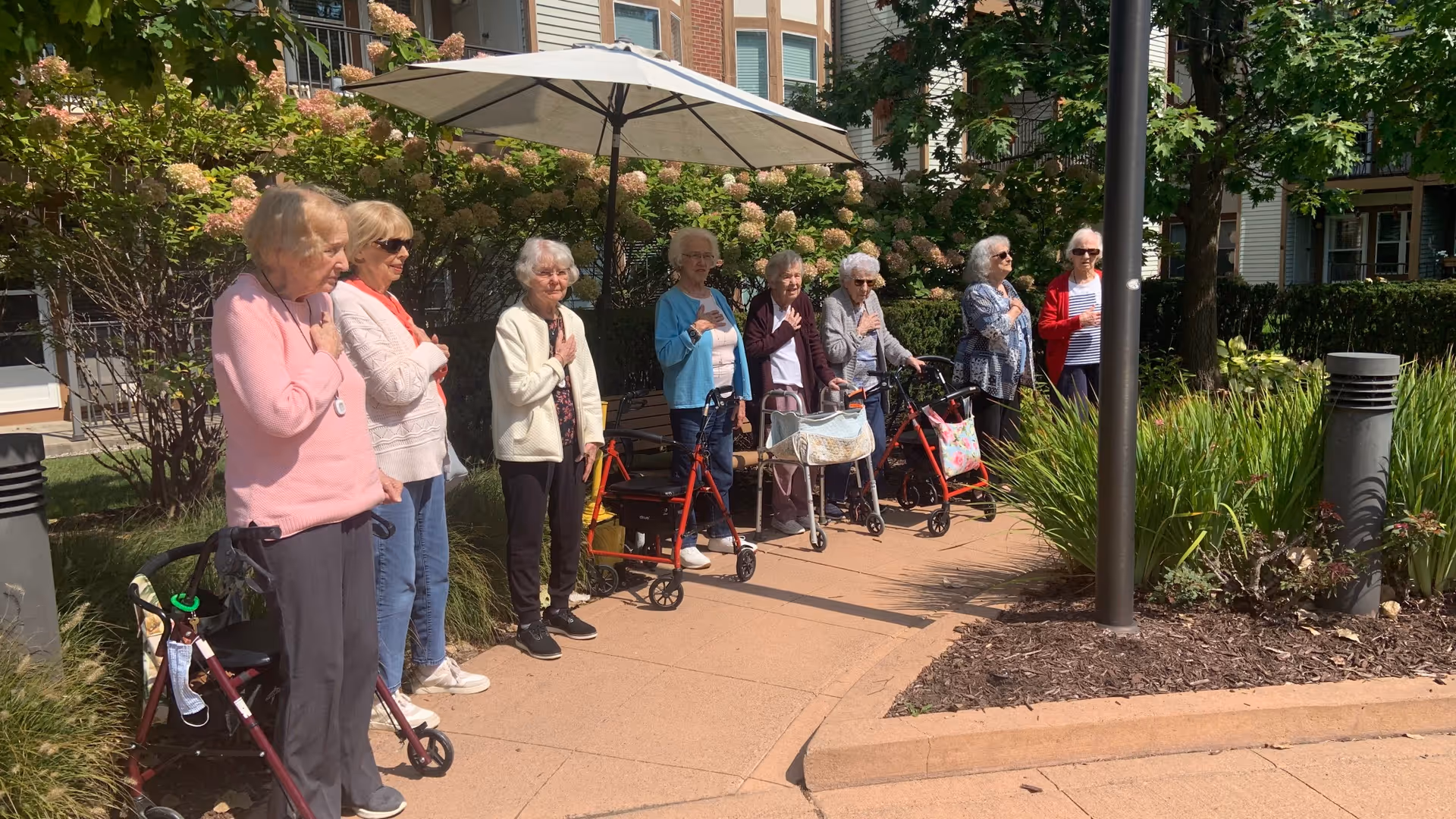 A group of elderly women standing outdoors on a paved pathway near a garden area with greenery and flowers. Some women are using walkers and all have their right hands placed over their hearts. They are standing in front of a building with windows and an umbrella providing shade.
