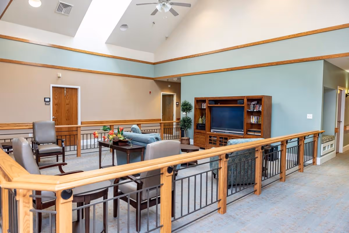 A spacious senior living facility common area with a high ceiling and ceiling fan. The room features comfortable seating including armchairs and sofas arranged around a wooden coffee table with a floral centerpiece. A wooden entertainment center with a TV and shelves is against a light blue wall. The area is bordered by a wooden and metal railing, and there are doors and a hallway visible in the background.