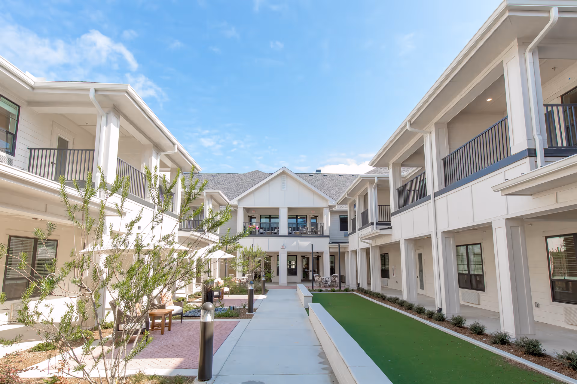 Outdoor courtyard area of a senior living facility with two-story white buildings on either side, balconies, a paved walkway, green lawn area, and some small trees and shrubs under a partly cloudy blue sky.