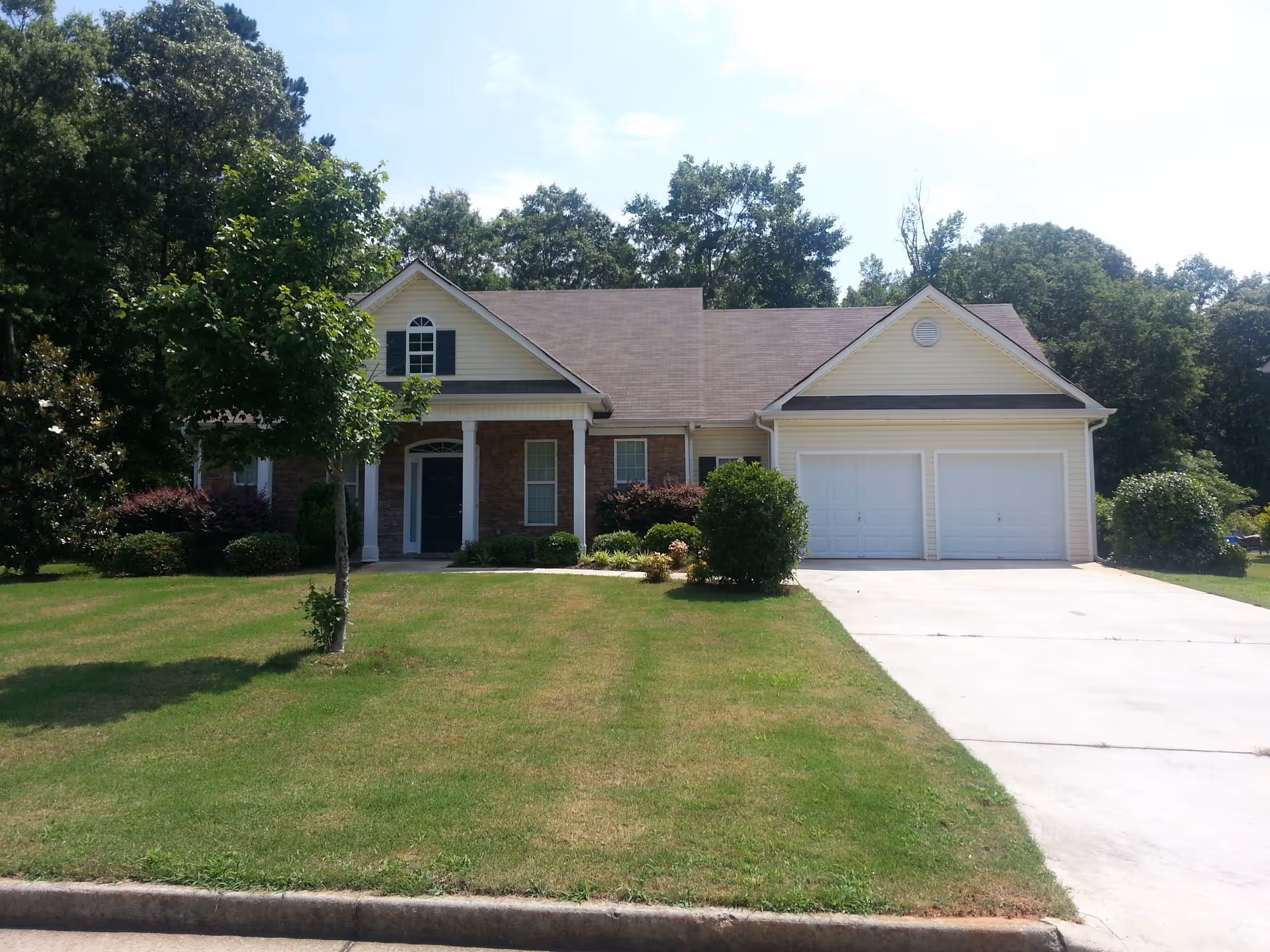 Front exterior view of a single-story house with a double garage, a driveway, a green lawn, and several bushes and trees under a clear sky.