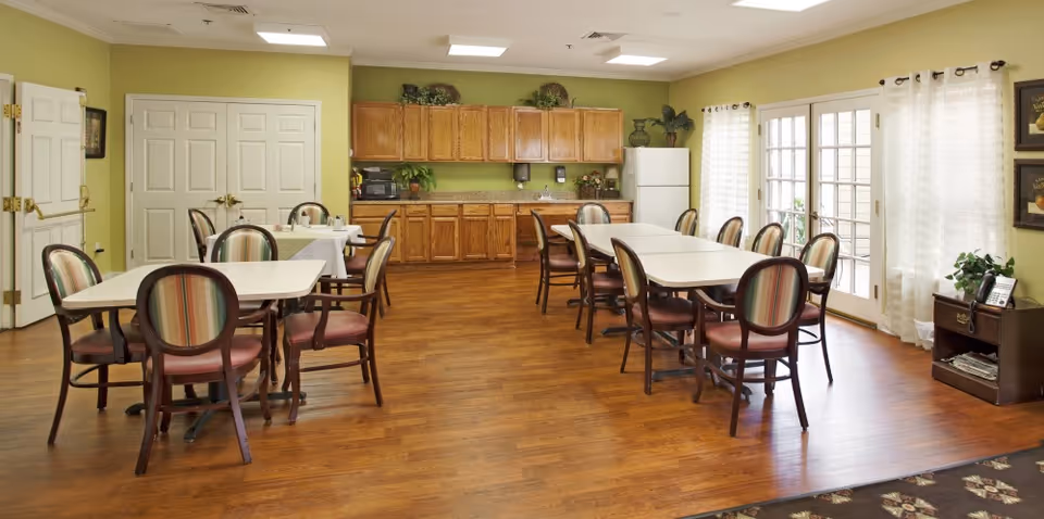 A dining room in a senior living facility with several tables and chairs arranged on a wooden floor. The room has light green walls, wooden cabinets, a white refrigerator, and a countertop with a microwave and plants. Large windows with sheer white curtains and a glass door allow natural light to enter the room.