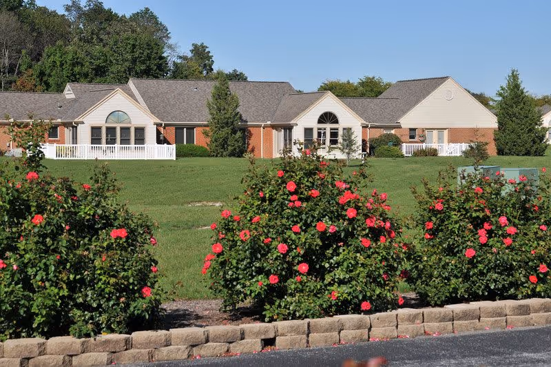 Single-story brick and siding senior living building set behind a lawn with a row of blooming red rose bushes in the foreground.