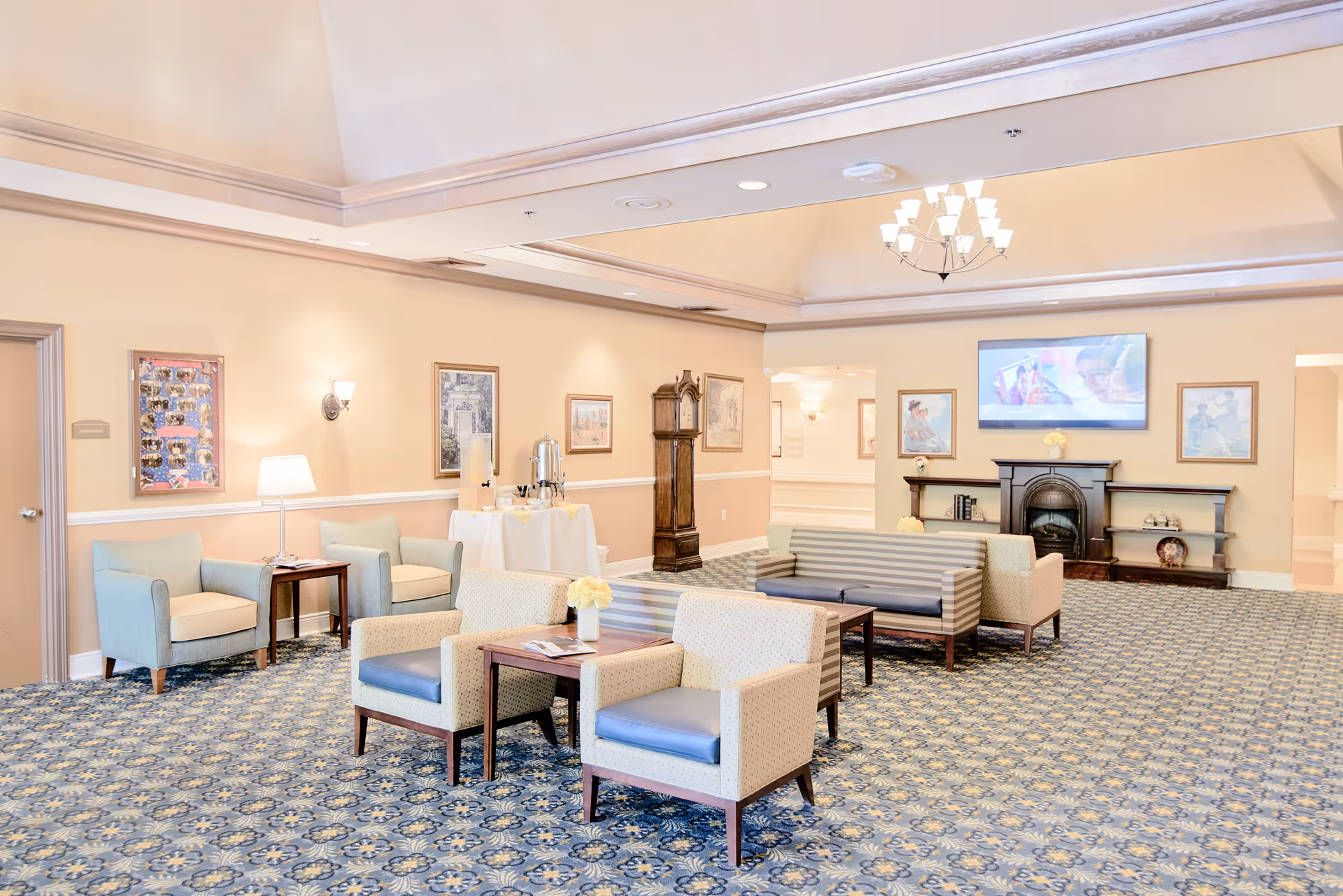 A spacious and well-lit living room area in a senior living facility with patterned carpet, several armchairs and sofas arranged around wooden coffee tables, a grandfather clock, framed artwork on the walls, a fireplace with a TV mounted above it, and a refreshment table with beverage dispensers.