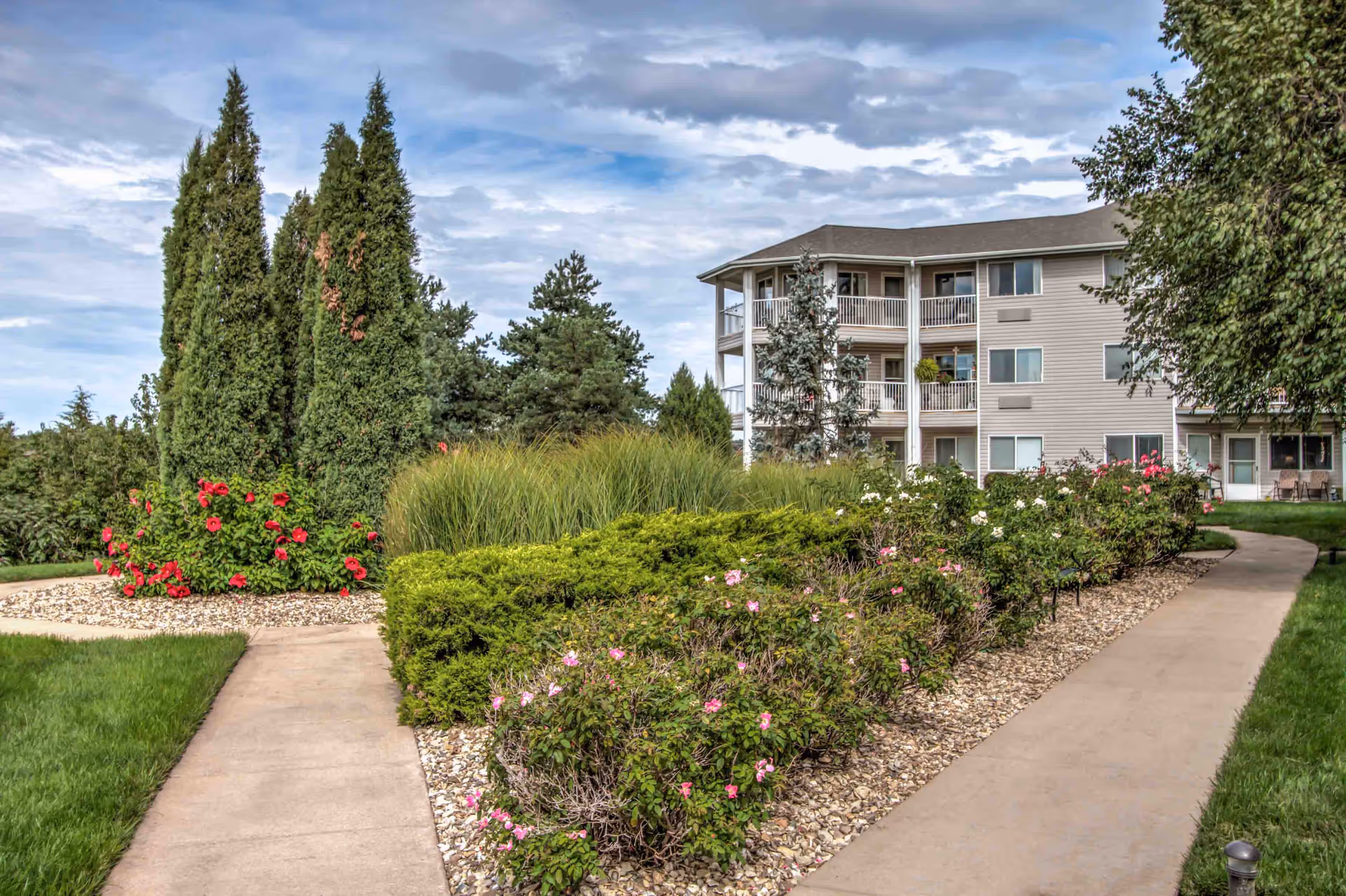 Landscaped walkway leading past flowerbeds to a three-story residential building with balconies under a cloudy sky.