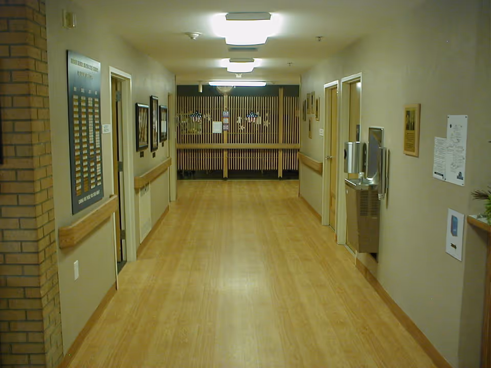 Well-lit interior hallway of a senior living facility with wood flooring, handrails, doors, and a water fountain.