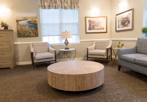 A cozy living room area with two striped armchairs, a gray sofa, a round wooden coffee table in the center, a small side table with a lamp between the armchairs, framed artwork on the walls, and a window with blinds and a floral valance.