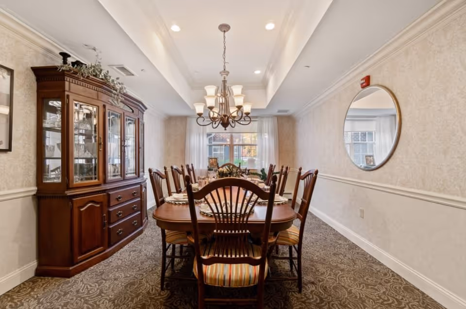 A formal dining room with a long wooden dining table surrounded by eight wooden chairs with striped cushions. A chandelier with multiple lights hangs above the table. On the left side, there is a large wooden china cabinet with glass doors displaying dishes. The walls are decorated with light-colored wallpaper and a round mirror hangs on the right wall. A window with sheer curtains is at the far end of the room, allowing natural light to enter.