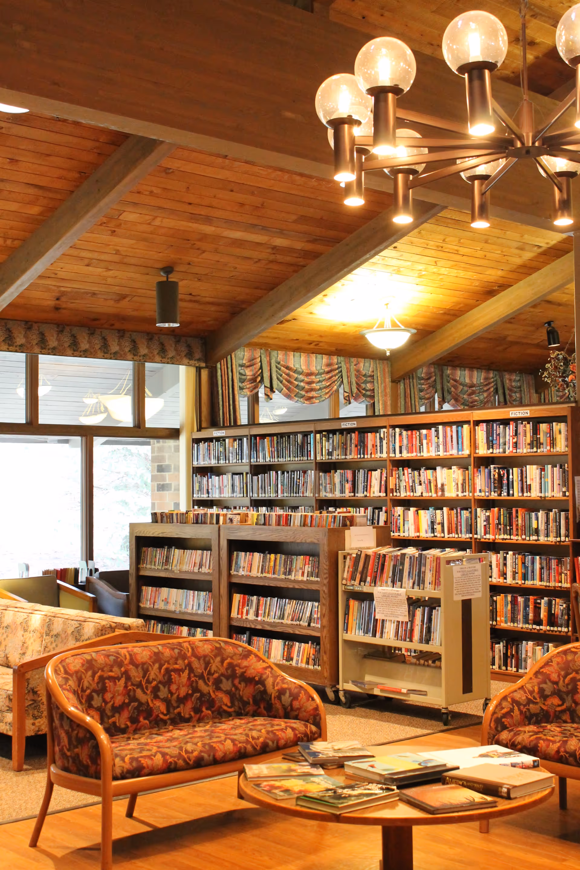 Cozy library room with wooden ceiling beams and warm lighting, featuring bookshelves filled with books labeled 'Fiction', patterned upholstered chairs, a round wooden table with books on it, and large windows with draped curtains.