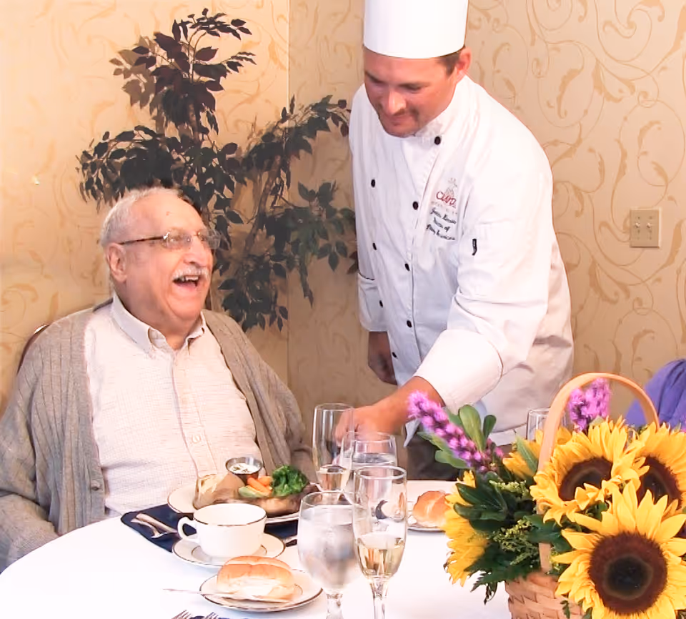 A smiling elderly man seated at a decorated dining table while a chef serves him and a basket of sunflowers sits nearby.