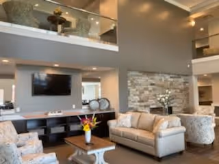 Spacious senior living lounge with sofas and armchairs around a coffee table, a wall-mounted TV, stone accent wall, and a double-height ceiling with a mezzanine.