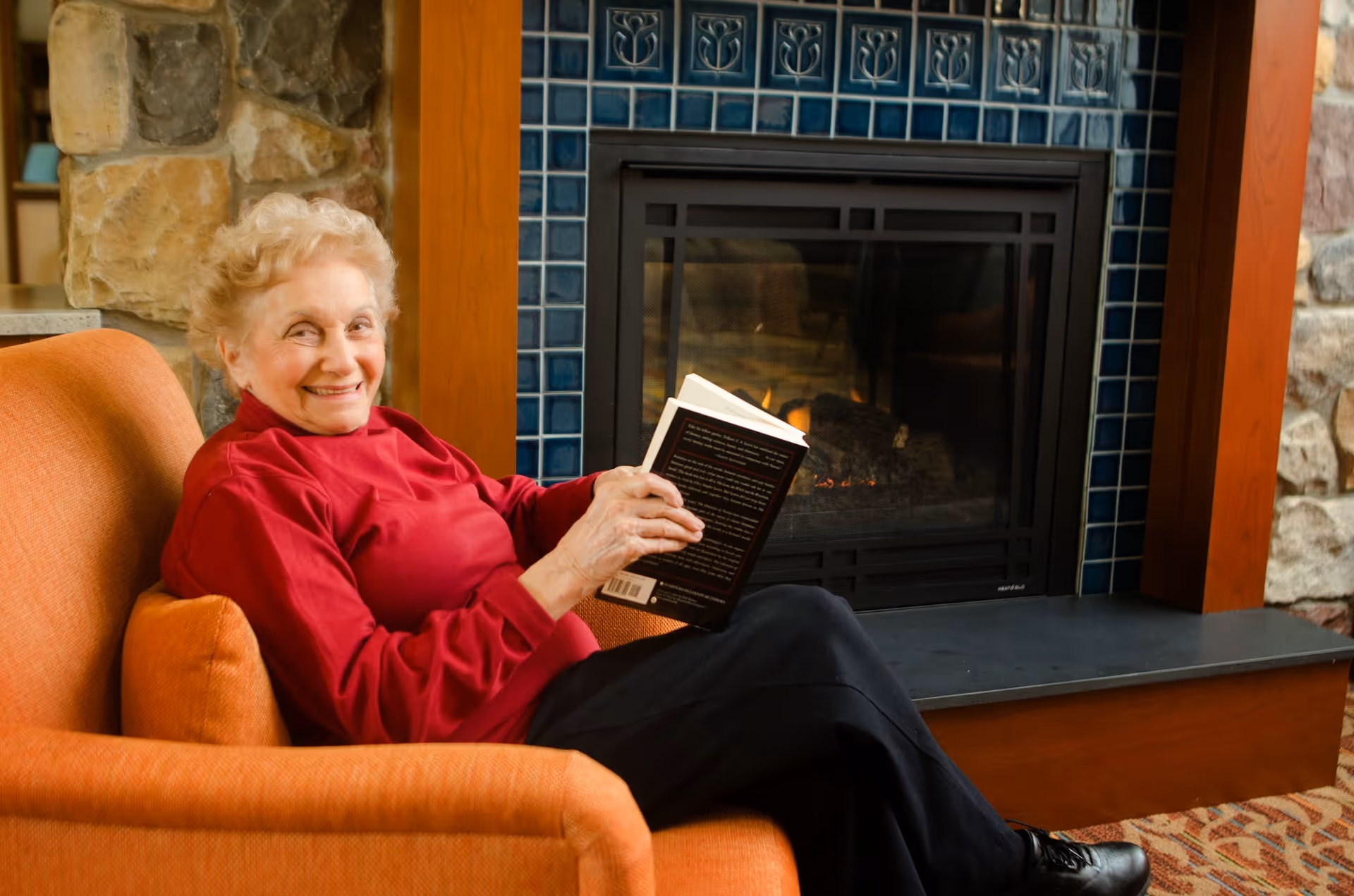 An elderly woman sits in an armchair by a fireplace smiling while holding an open book.