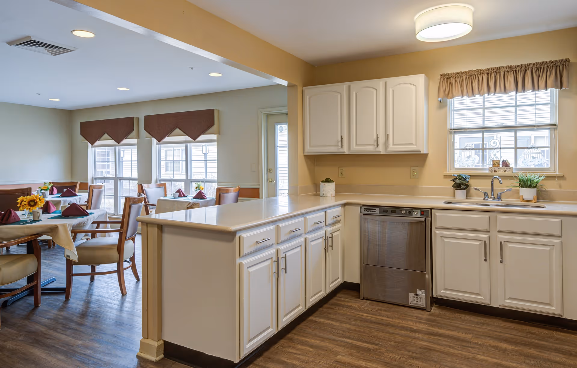 A bright kitchen area with white cabinets, a stainless steel dishwasher, and a sink under a window with beige curtains. Adjacent to the kitchen is a dining area with several tables covered with beige tablecloths, each set with maroon napkins and small flower arrangements. The floor is wood, and the walls are painted a light beige color.