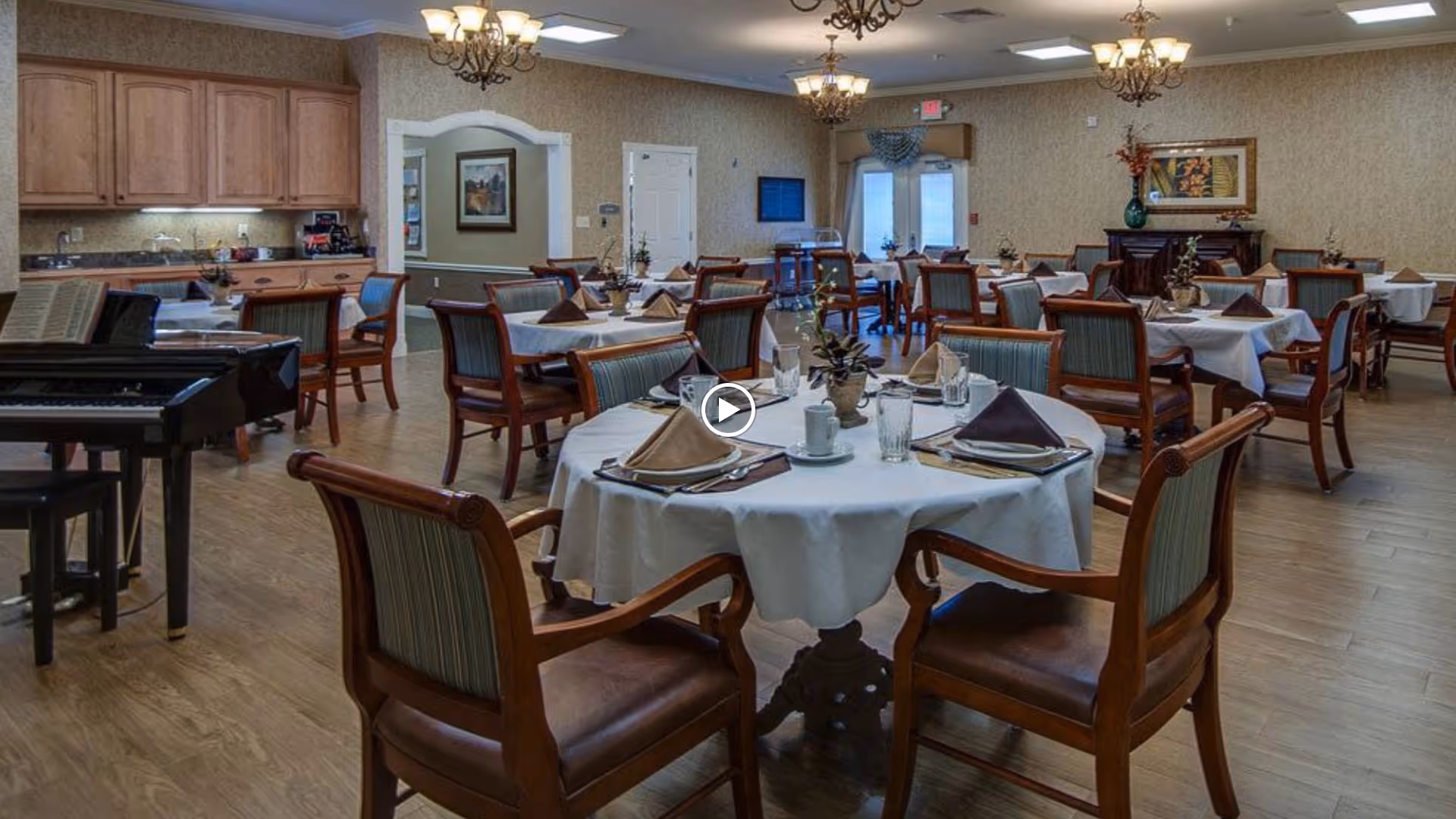 A dining room in a senior living facility with multiple round tables covered with white tablecloths, each set with folded napkins, plates, cups, and glasses. Wooden chairs with cushioned seats surround the tables. The room has wooden flooring, chandeliers hanging from the ceiling, and a piano on the left side near a kitchen area with wooden cabinets.