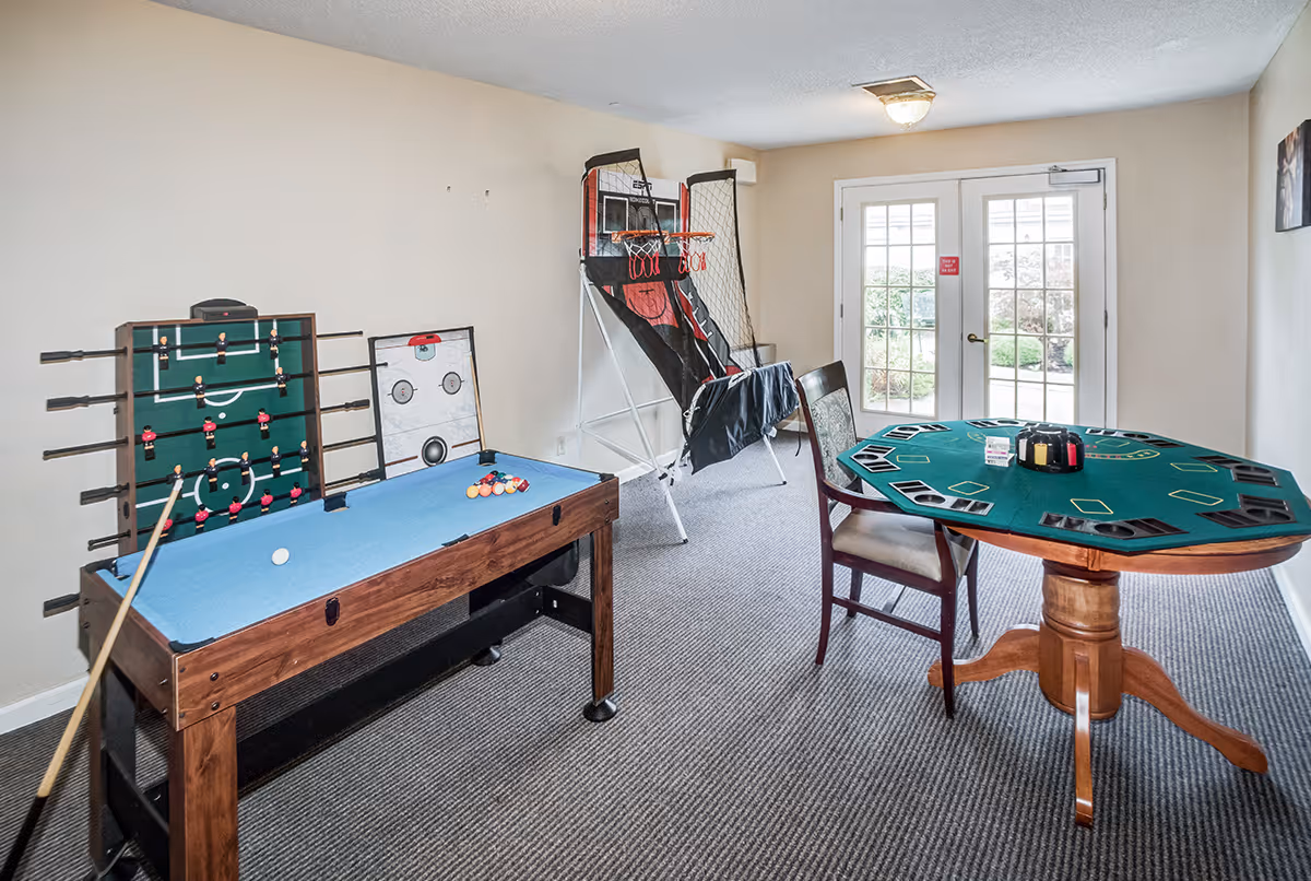A game room with a pool table, foosball table, tabletop hockey game, basketball arcade game, and a round card table with chairs. French doors lead outside to a garden area.