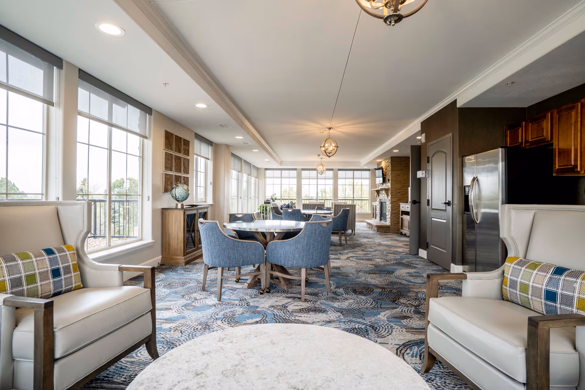 A bright and spacious common area in an assisted living facility featuring large windows with views outside, multiple round tables with blue upholstered chairs, two white armchairs with plaid cushions, a patterned carpet, and a stone fireplace in the background. The room has a modern and comfortable design with overhead lighting fixtures.