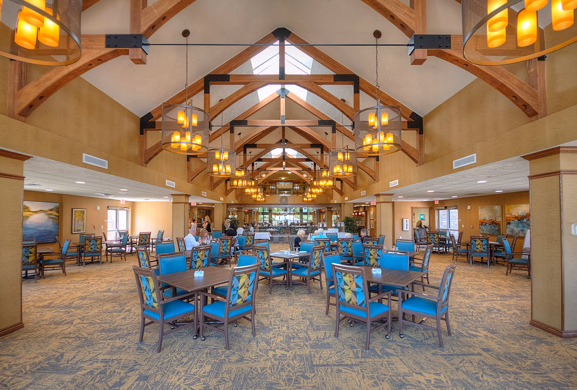 Spacious dining room with high vaulted ceilings featuring wooden beams and multiple hanging light fixtures. The room has several tables with blue cushioned chairs arranged neatly, and a few people are seated and standing in the background near the serving area. The walls are painted in warm tones with large windows allowing natural light to enter.