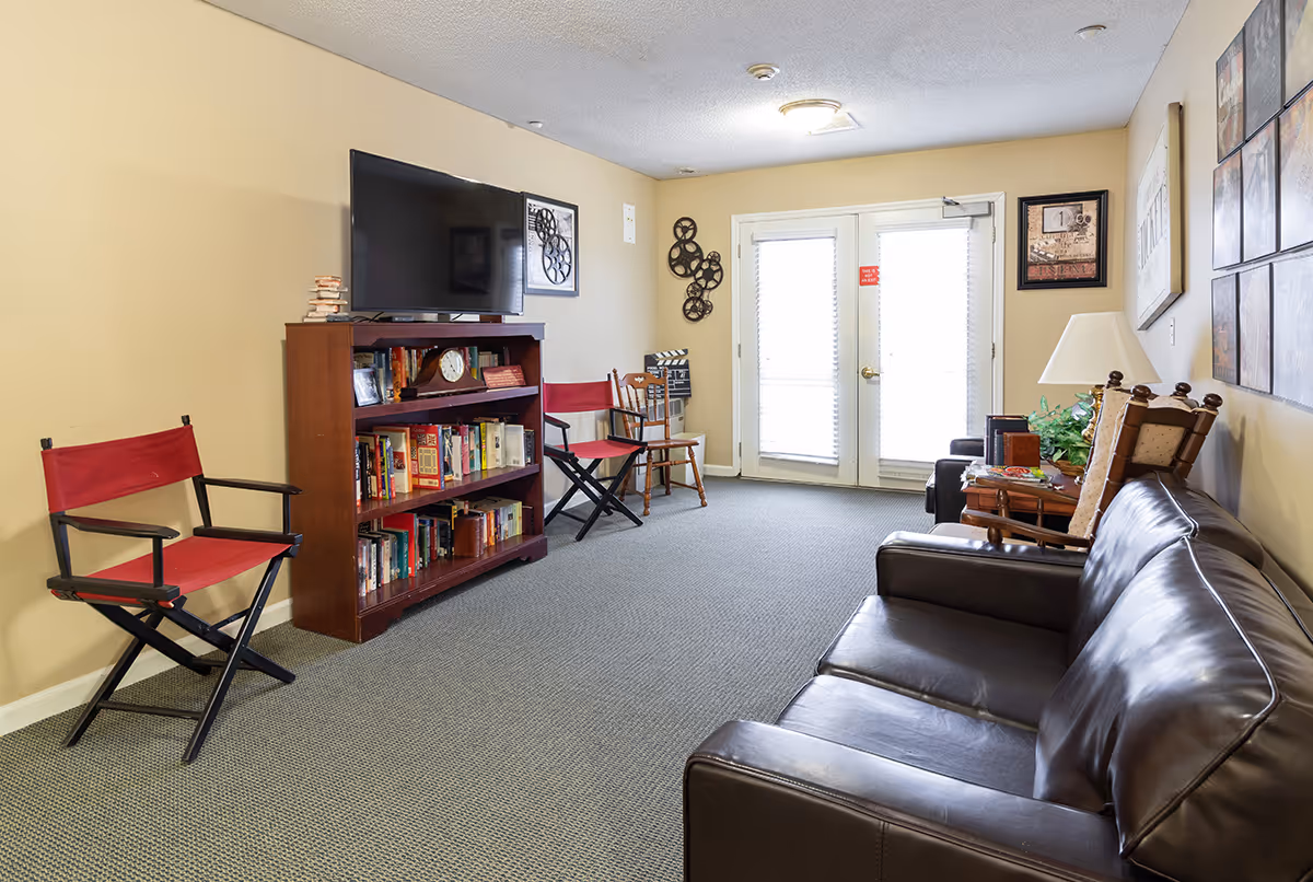 A cozy living room area with a dark brown leather sofa, two red director-style chairs, and a wooden chair. There is a wooden bookshelf filled with books and a clock, with a flat-screen TV mounted above it. The room has beige walls adorned with framed pictures and decorative wall art. A small wooden side table with a lamp and a plant is next to the sofa. Double doors with blinds allow natural light into the room.