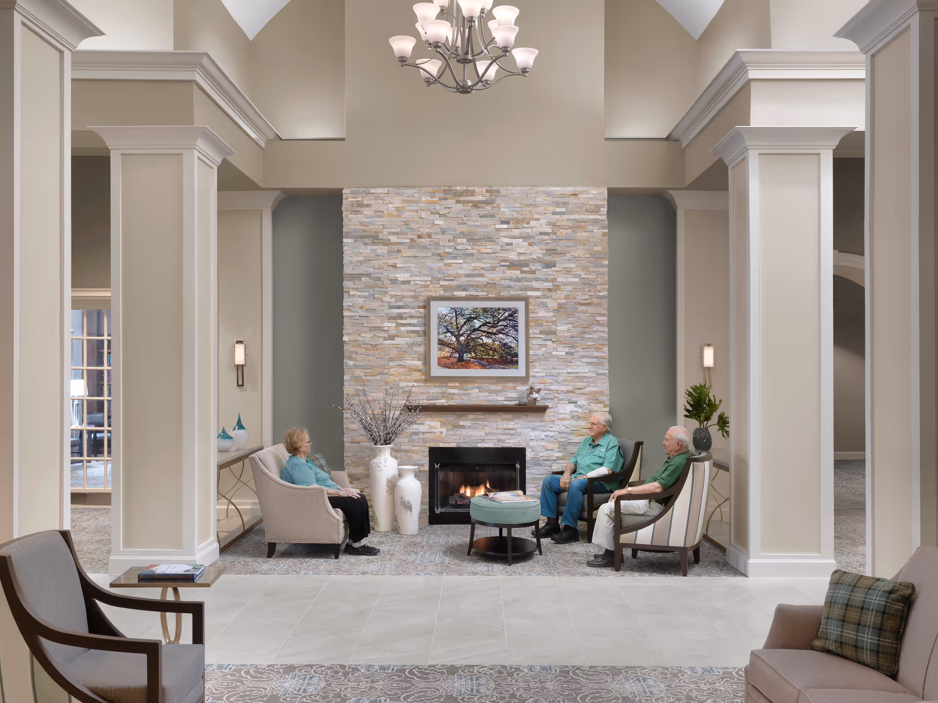 A cozy living room area in Buckner Parkway Place featuring a stone fireplace with a framed picture of a tree above it. Three elderly people are seated around the fireplace on comfortable chairs. The room has tall ceilings with crown molding, a chandelier, decorative vases, and soft lighting.