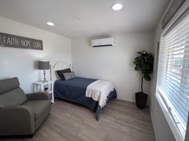 A small, neatly arranged bedroom with a single bed covered in a dark blue quilt and a white throw blanket. Next to the bed is a white nightstand with a lamp and a small plant. There is a gray armchair in the corner, a potted plant near the window, and a wall-mounted air conditioner above the bed. The walls are white, and there is a sign on the wall that reads 'FAITH · HOPE · LOVE'. Light streams in through a window with white blinds.
