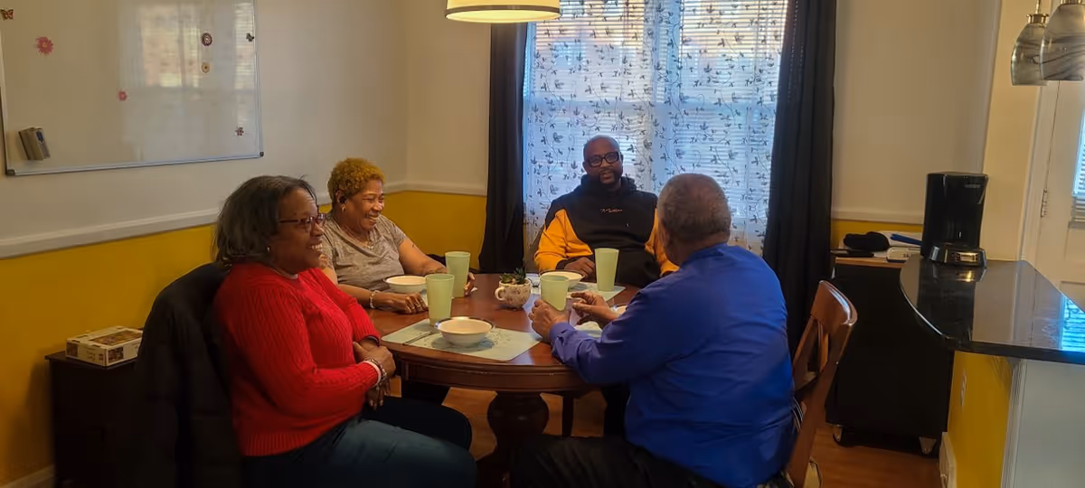 Four adults sitting around a round wooden dining table in a cozy room with yellow and white walls. They are engaged in conversation and smiling, with cups and bowls on the table. A window with patterned curtains and a hanging light fixture are visible in the background.