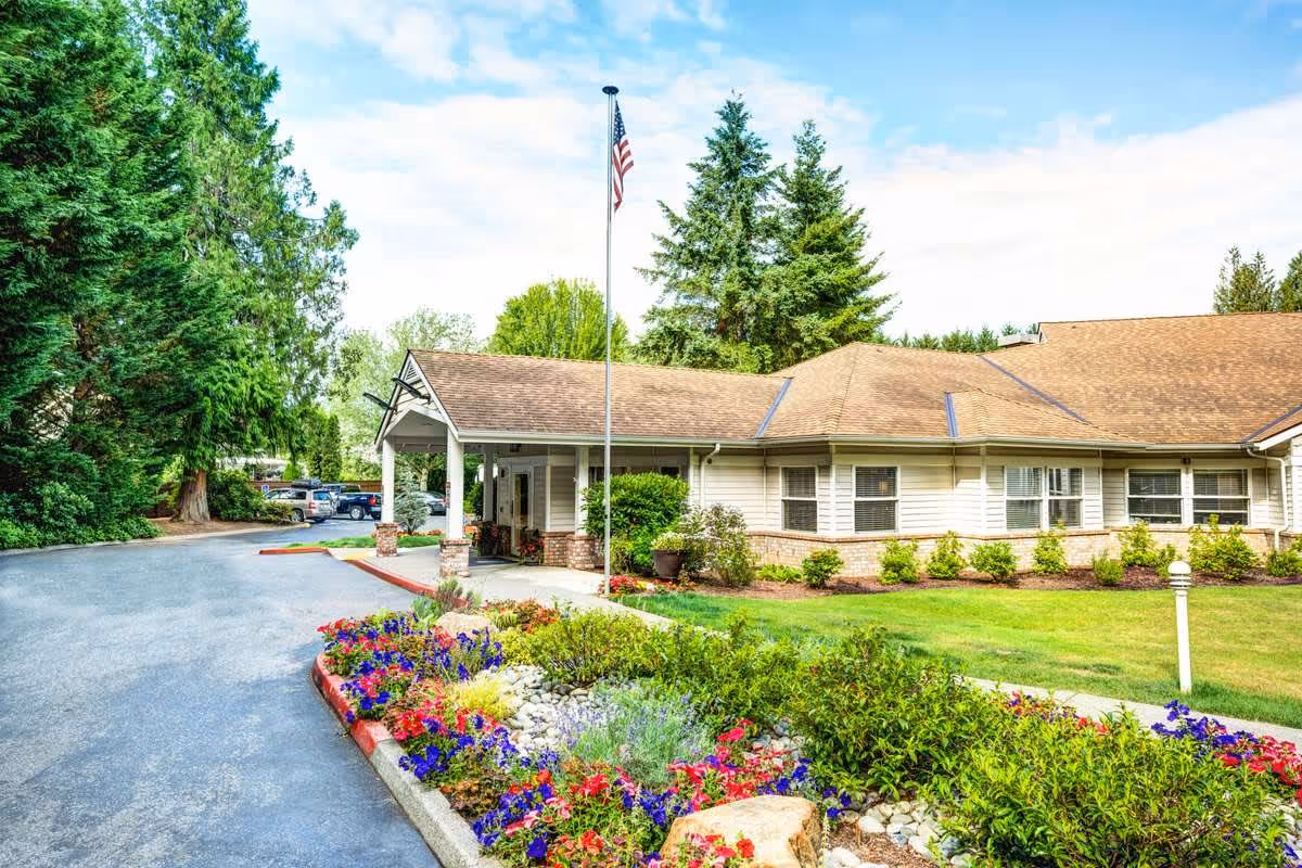 Exterior view of a single-story building with a covered entrance, surrounded by a well-maintained garden with colorful flowers and green shrubs. An American flag is displayed on a flagpole near the entrance, and tall trees are visible in the background under a partly cloudy sky.