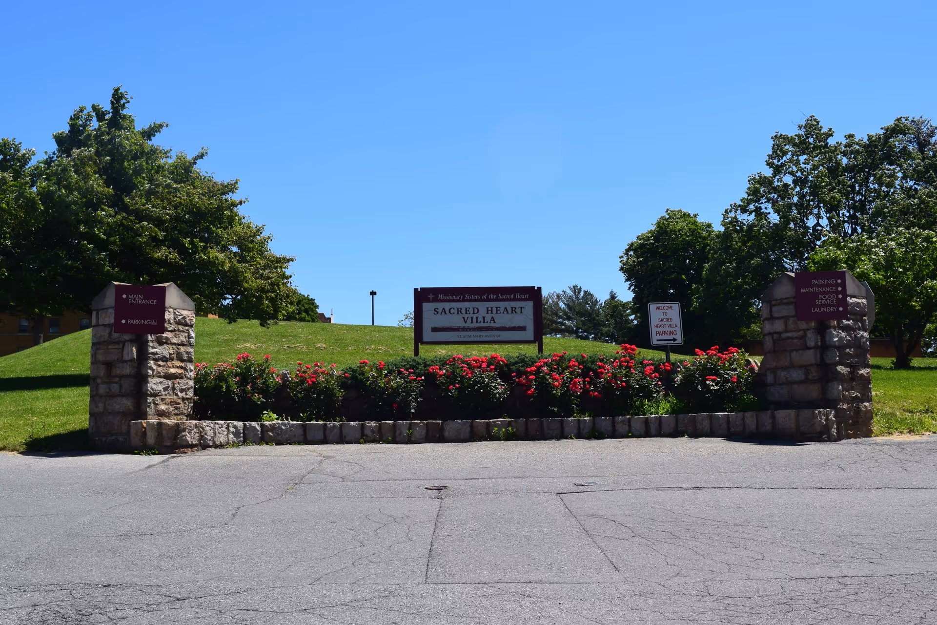 Entrance sign reading 'Sacred Heart Villa' set between stone pillars with roses, lawn, and trees under a clear blue sky.