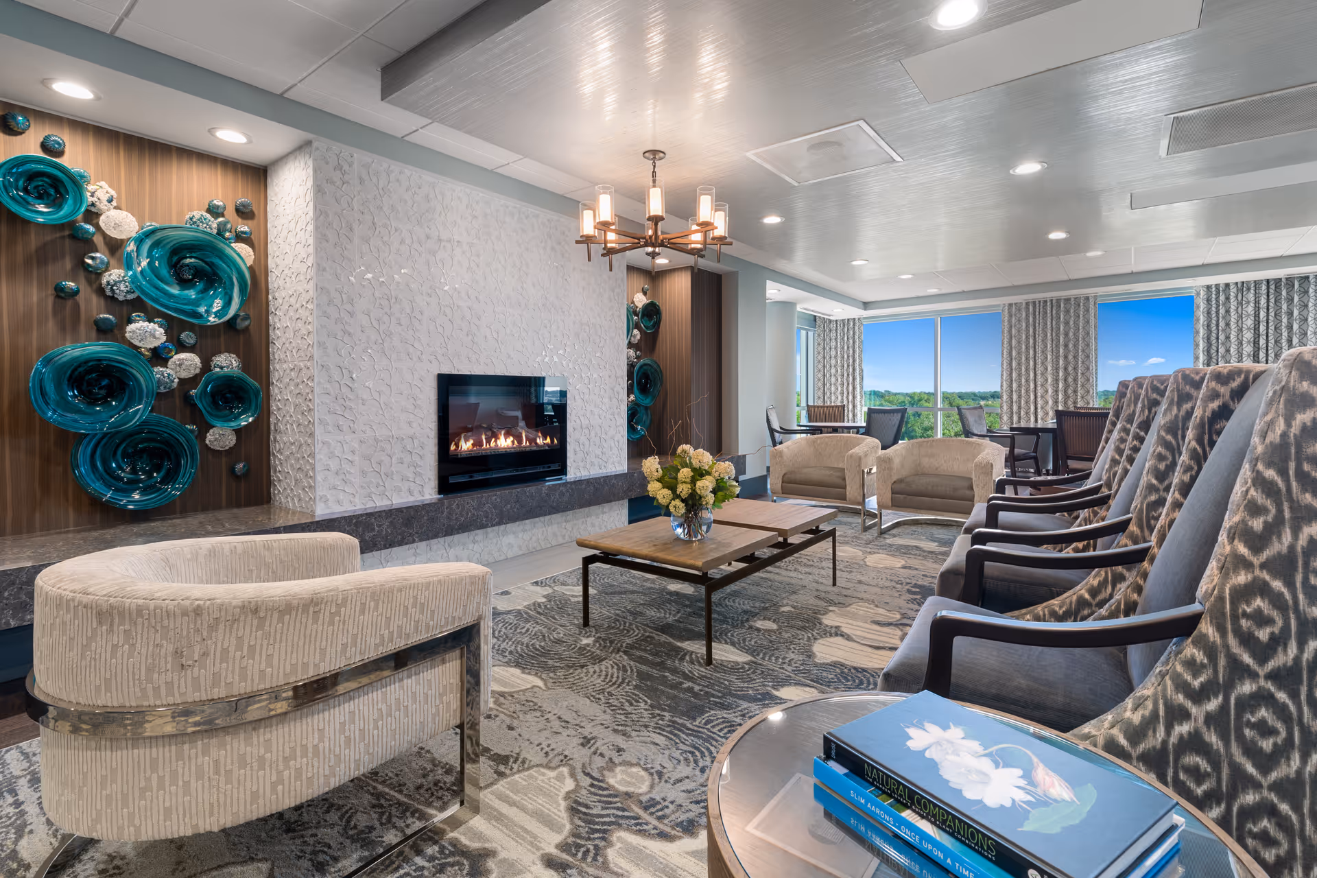 A modern living room with a fireplace set in a white textured wall, surrounded by decorative teal and white wall art. The room features a variety of seating including patterned armchairs, beige chairs, and a wooden coffee table with a vase of flowers. Large windows with patterned curtains allow natural light to fill the space, and a chandelier hangs from the ceiling.