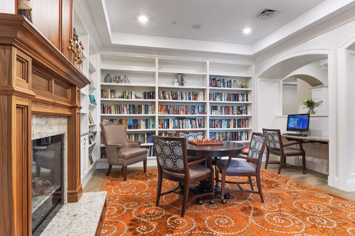 A cozy library and sitting area in a senior living facility featuring a wooden fireplace on the left, white built-in bookshelves filled with books, a round table with a chess set and four chairs, an armchair, and a computer workstation with a desktop computer on the right. The floor is covered with an orange patterned carpet.