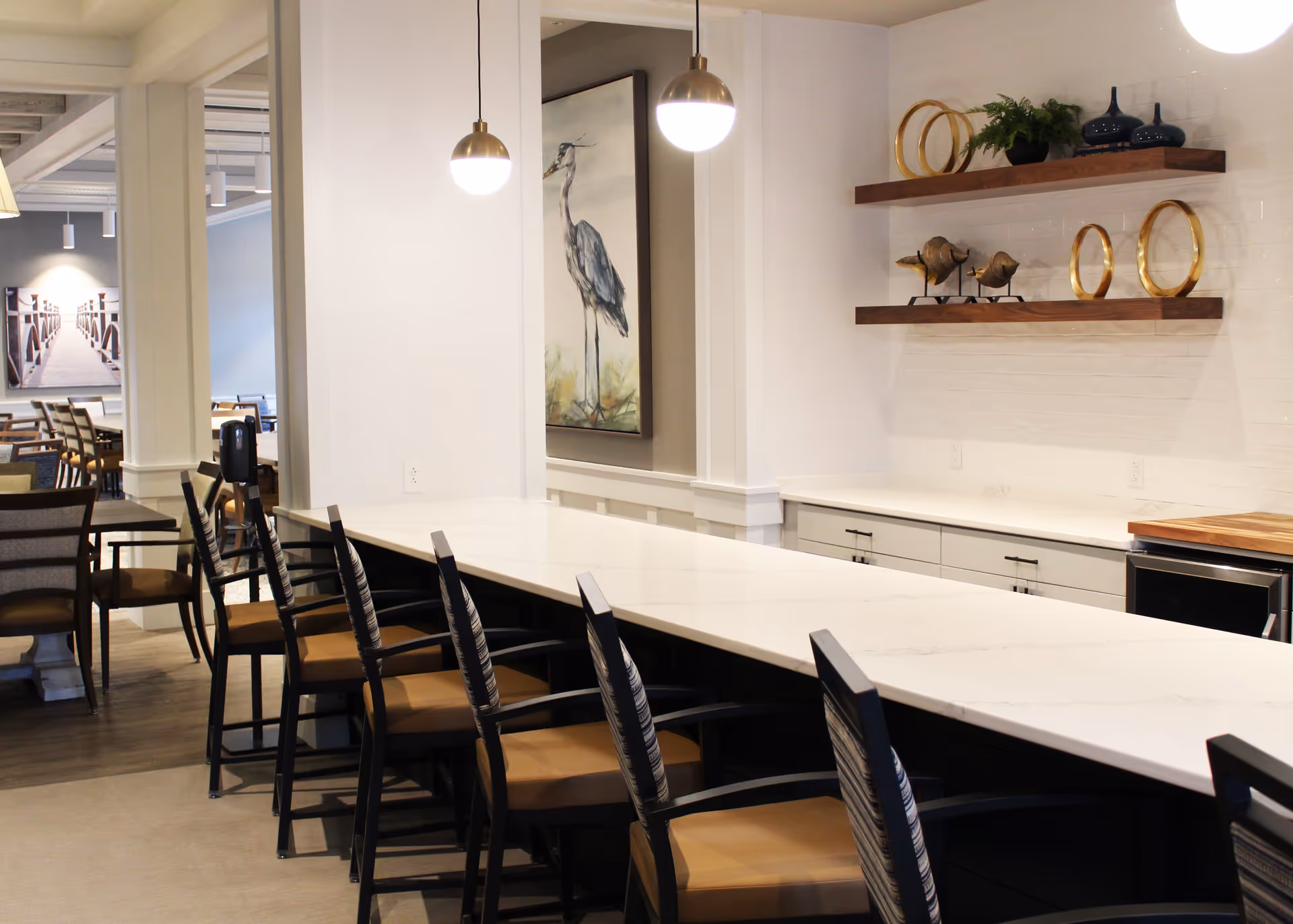 Interior view of a modern dining area with a long white countertop and several chairs with armrests. Two pendant lights hang above the countertop. On the right wall, there are two wooden floating shelves decorated with plants, circular gold ornaments, and bird sculptures. In the background, there are additional dining tables and chairs, and a large framed artwork of a bird on the wall.