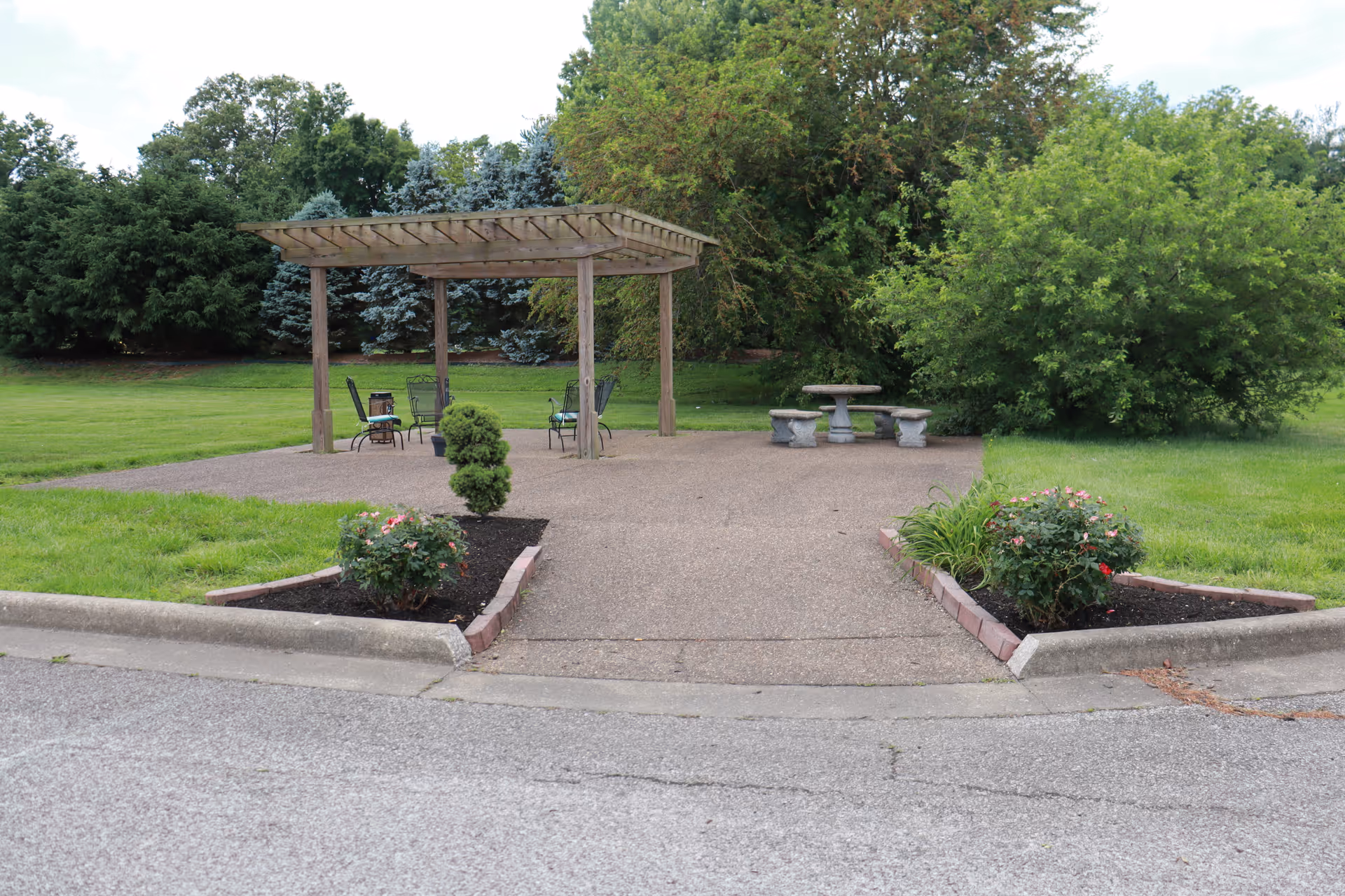 Outdoor seating area with a wooden pergola covering several metal chairs and a small table. There is a concrete picnic table with benches nearby, surrounded by green grass, bushes, and trees.