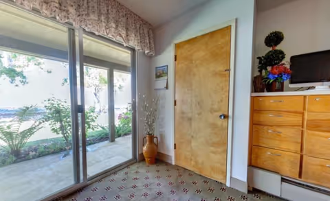 Interior view of a room with a sliding glass door leading to an outdoor garden area. The room has a wooden door, a wooden dresser with a small TV and decorative plants on top, and a patterned carpet. A floral valance is above the sliding door.
