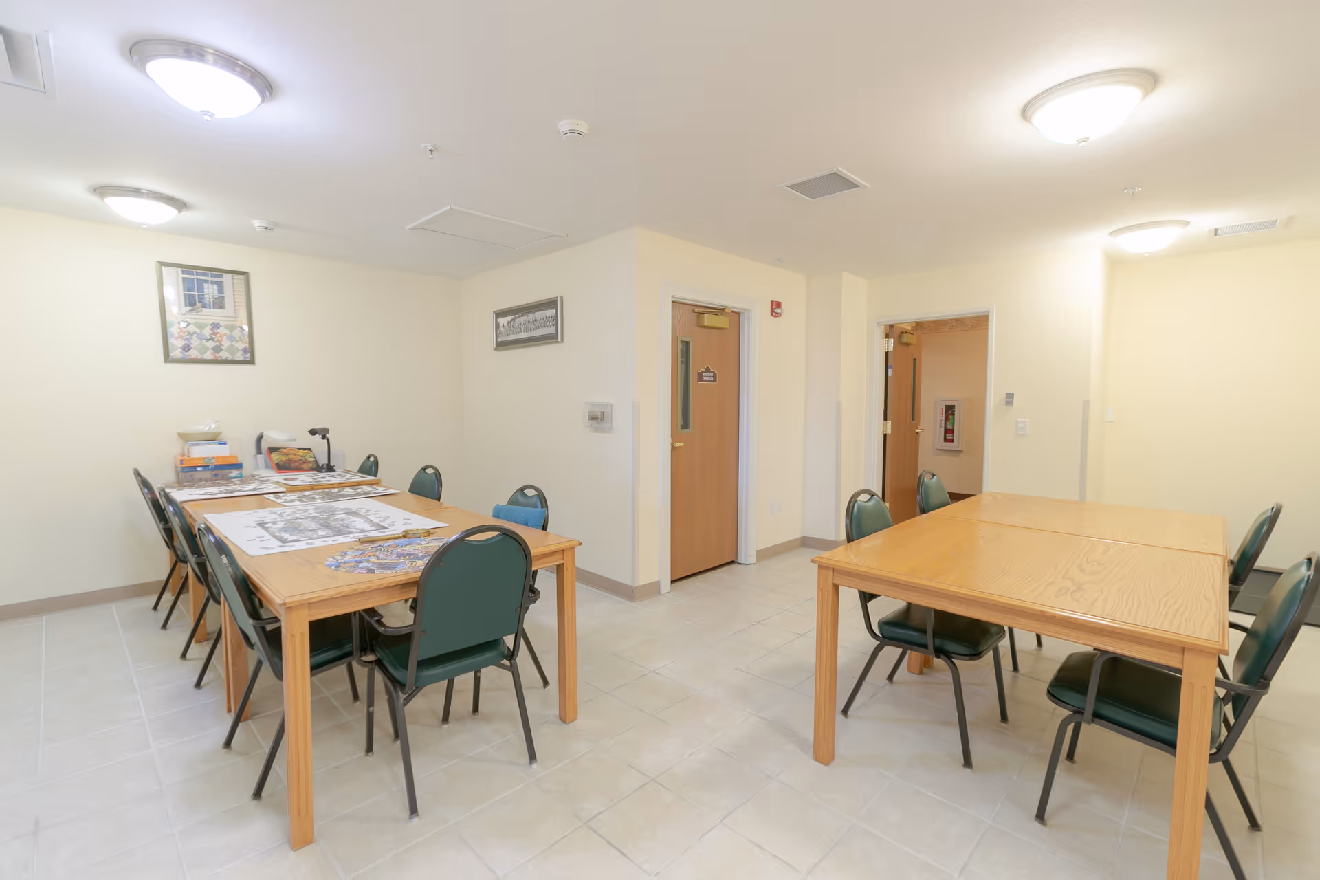 Well-lit common room with two wooden tables surrounded by green chairs and light yellow walls.