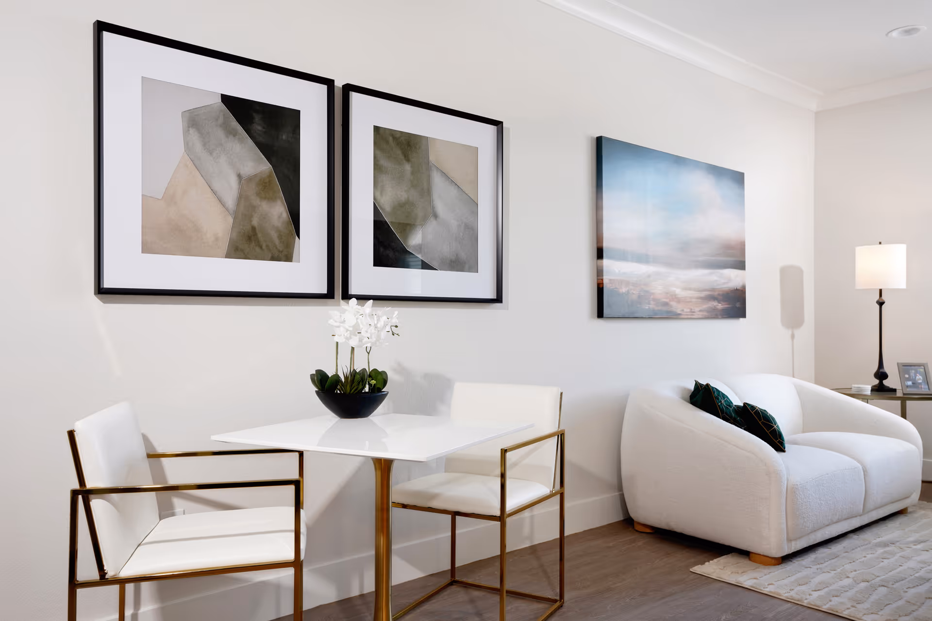 A modern living area with a small white table and two white chairs with gold frames. On the table is a black vase with white flowers. Behind the table are two abstract framed artworks on the wall. To the right is a white cushioned loveseat with two dark green pillows, a side table with a lamp, and a framed photo. The floor is wooden, and the walls are light-colored.