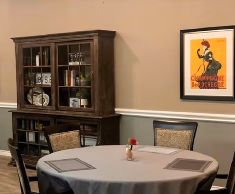 A dining area with a round table covered with a gray tablecloth and four chairs around it. A small vase with a red flower is placed in the center of the table. Behind the table is a dark wooden cabinet with glass doors displaying books, plates, and decorative items. On the beige wall above the chair is a framed vintage-style poster with the text 'CHAMPAGNE DE ROCHECRE'.