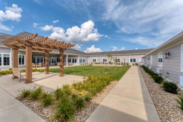 Outdoor courtyard area at Cedarhurst Senior Living of Springfield featuring a wooden pergola with seating underneath, surrounded by a well-maintained lawn, flower beds, and paved walkways. The building exterior is light-colored with multiple windows under a partly cloudy blue sky.