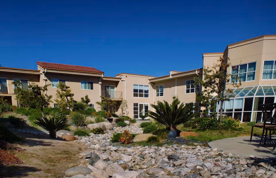 Outdoor garden area at Nikkei Senior Gardens featuring a dry rock creek bed surrounded by various plants and shrubs, with a beige two-story building in the background under a clear blue sky.