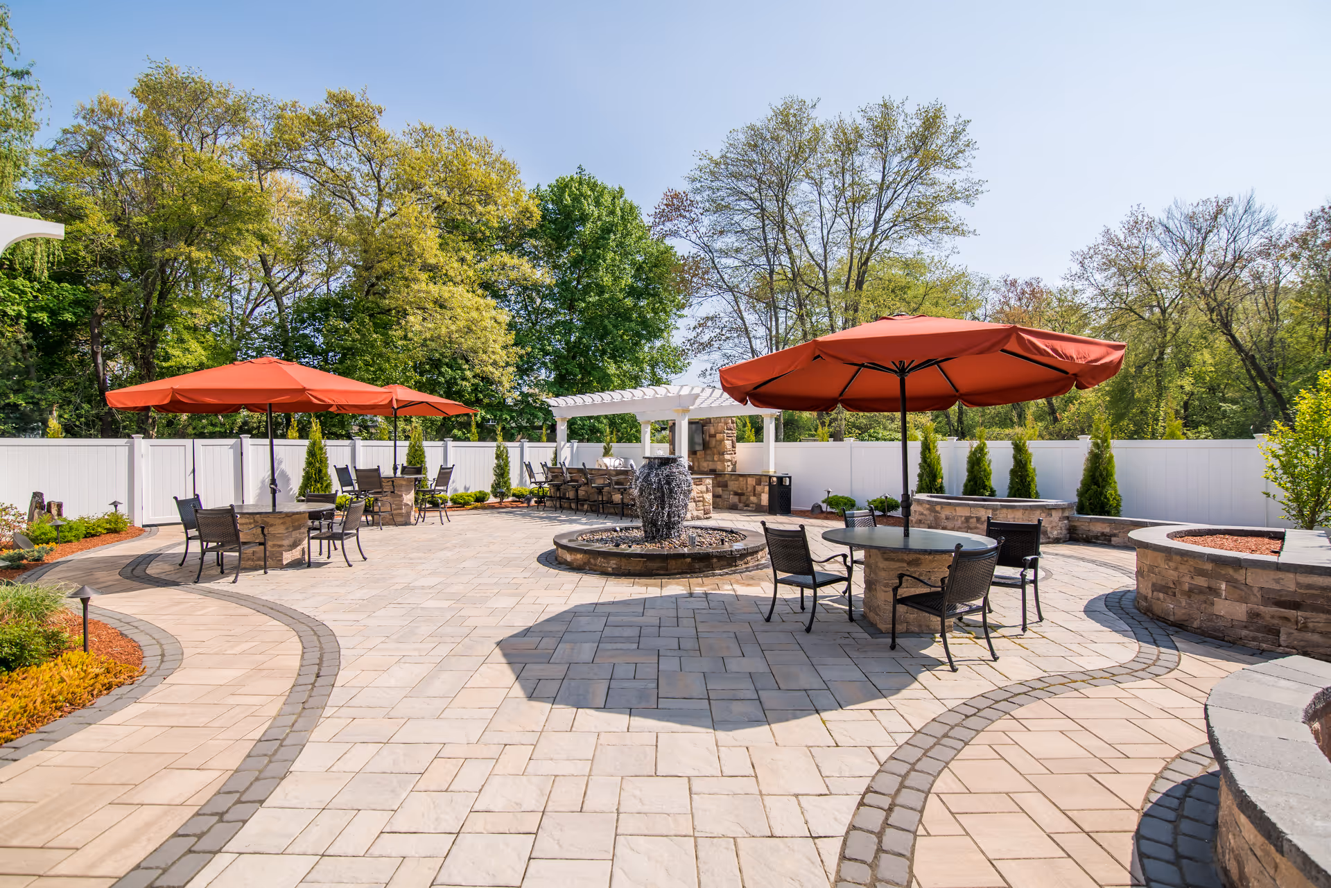 Outdoor patio area with stone-paved flooring, round tables with red umbrellas, black chairs, a central water fountain, and a white fence surrounding the space. Trees and greenery are visible in the background under a clear blue sky.