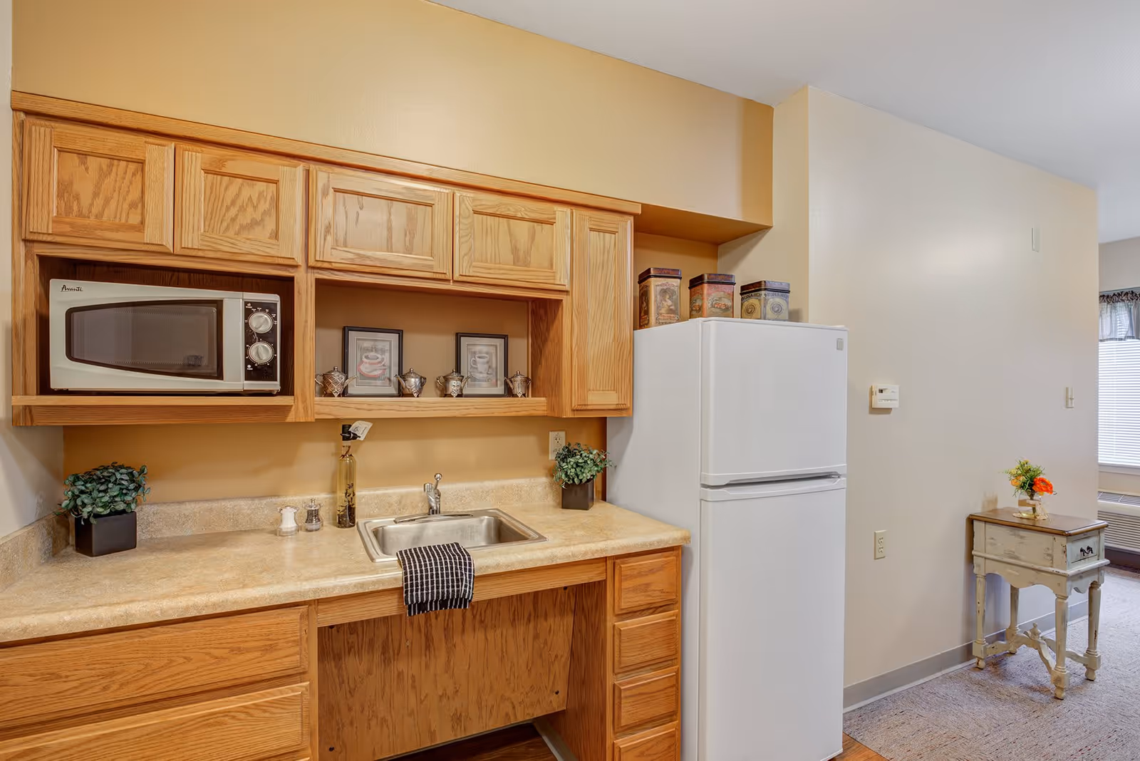A small kitchen area with wooden cabinets, a countertop with a sink, a microwave oven, and a white refrigerator. There are decorative items such as framed pictures, small plants, and tins on the countertop and above the refrigerator. To the right, there is a small vintage-style table with a flower vase on top, and a window with blinds in the background.
