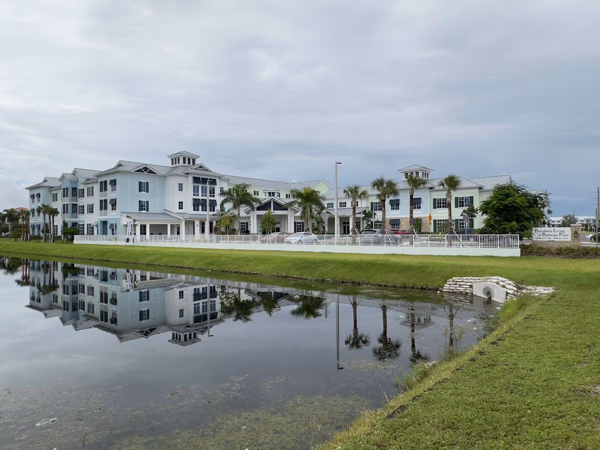 Front exterior of a multi-story senior living building reflected in a pond with palm trees and a grassy foreground.