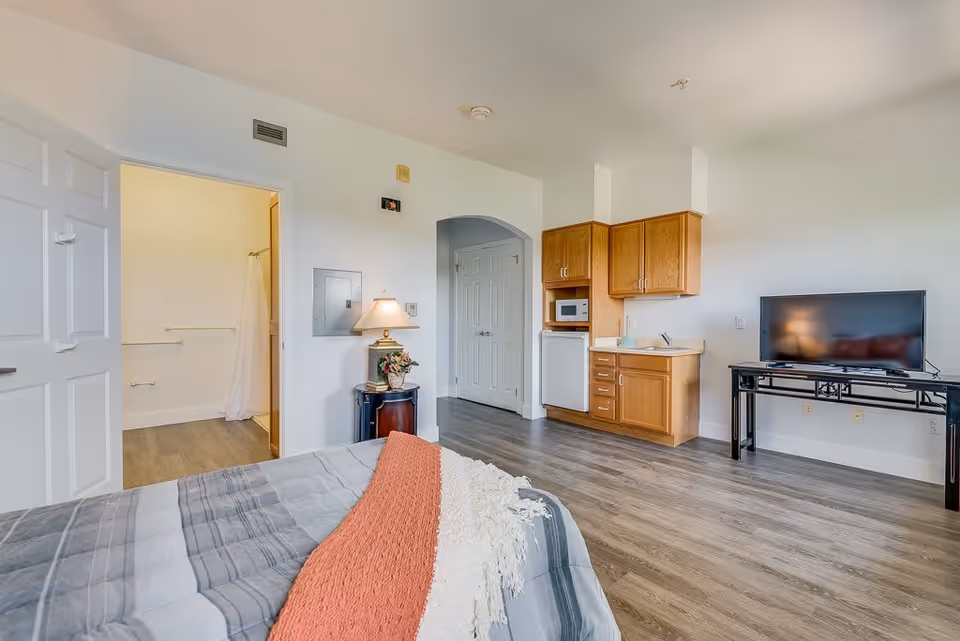 Interior view of a senior living facility room featuring a bed with a gray and orange blanket, a small kitchenette with wooden cabinets, a microwave, and a mini fridge. There is a TV on a black stand against the wall, a lamp on a small round table with a flower arrangement, and an open door leading to a bathroom with a shower curtain visible.