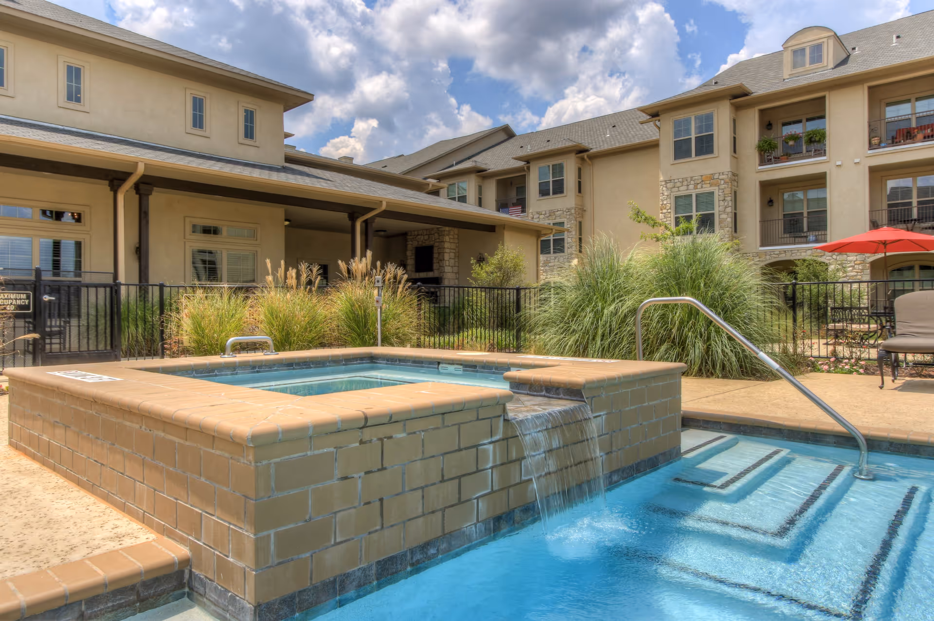 Outdoor pool area at Arabella of Longview Independent Living featuring a raised hot tub with water flowing into the pool, surrounded by beige brickwork. The background shows a multi-story residential building with balconies, windows, and some greenery, under a partly cloudy sky.