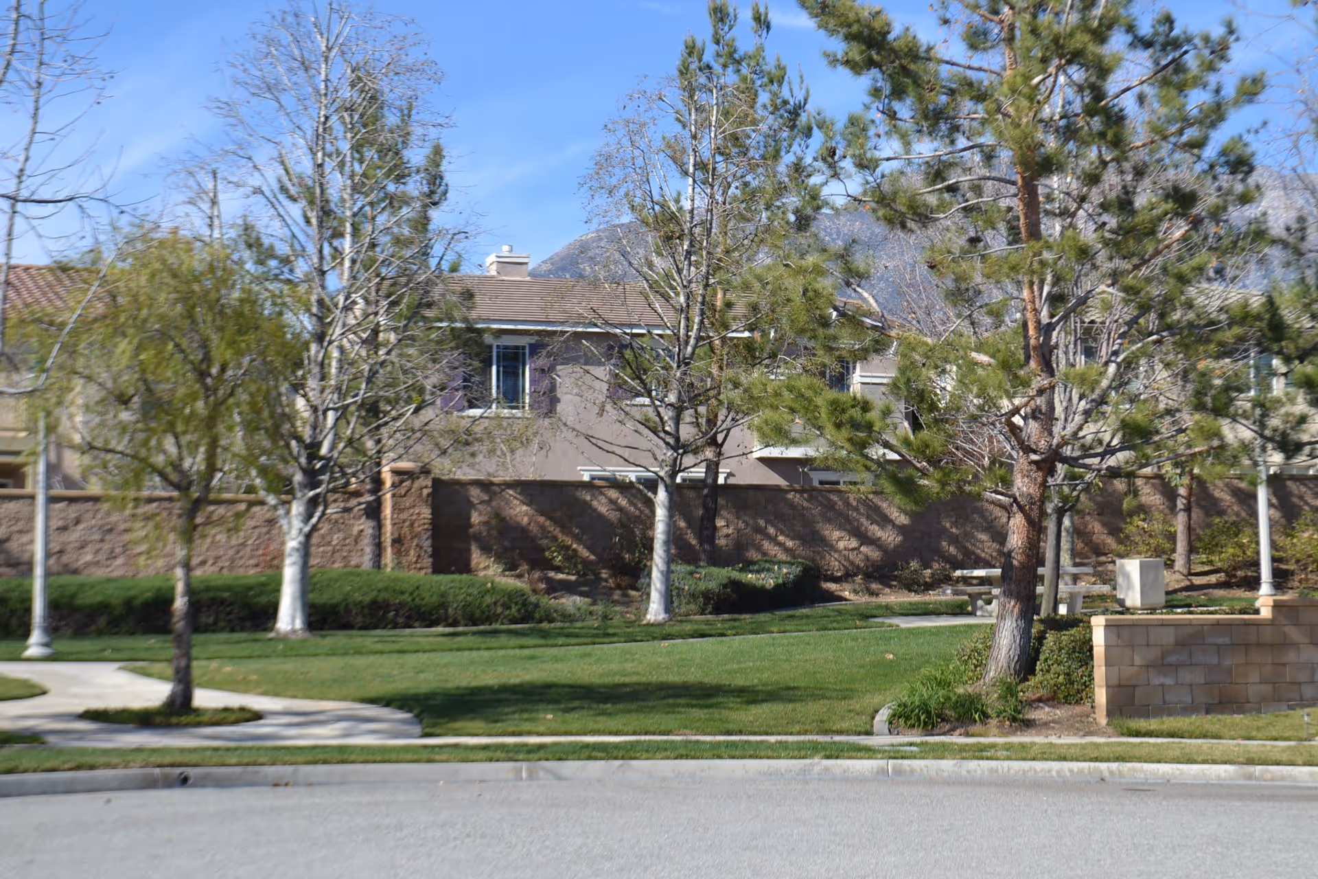 A landscaped outdoor area with green grass, several trees, a concrete sidewalk, and a stone wall. In the background, there is a residential building with windows and a tiled roof, and mountains under a clear blue sky.