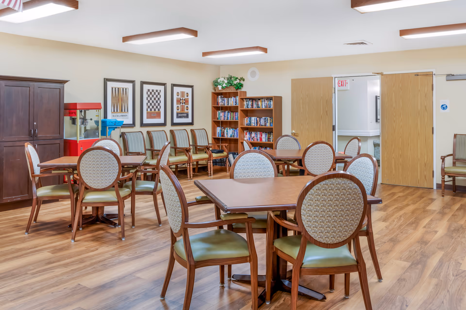 Well-lit communal dining/activity room with multiple tables and chairs, a popcorn machine, and a bookshelf.