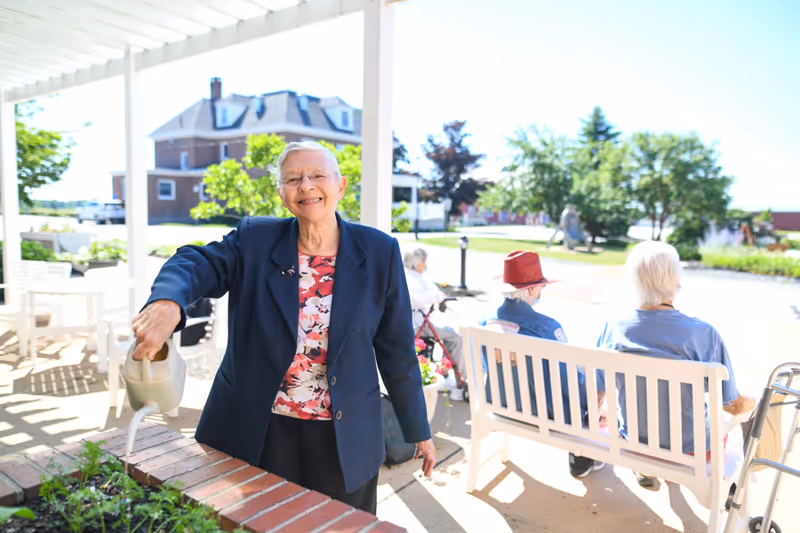 An elderly woman smiling and watering plants in a garden area outside a senior living facility. In the background, two other elderly individuals are seated on a white bench under a covered patio, with greenery and a building visible further behind them.