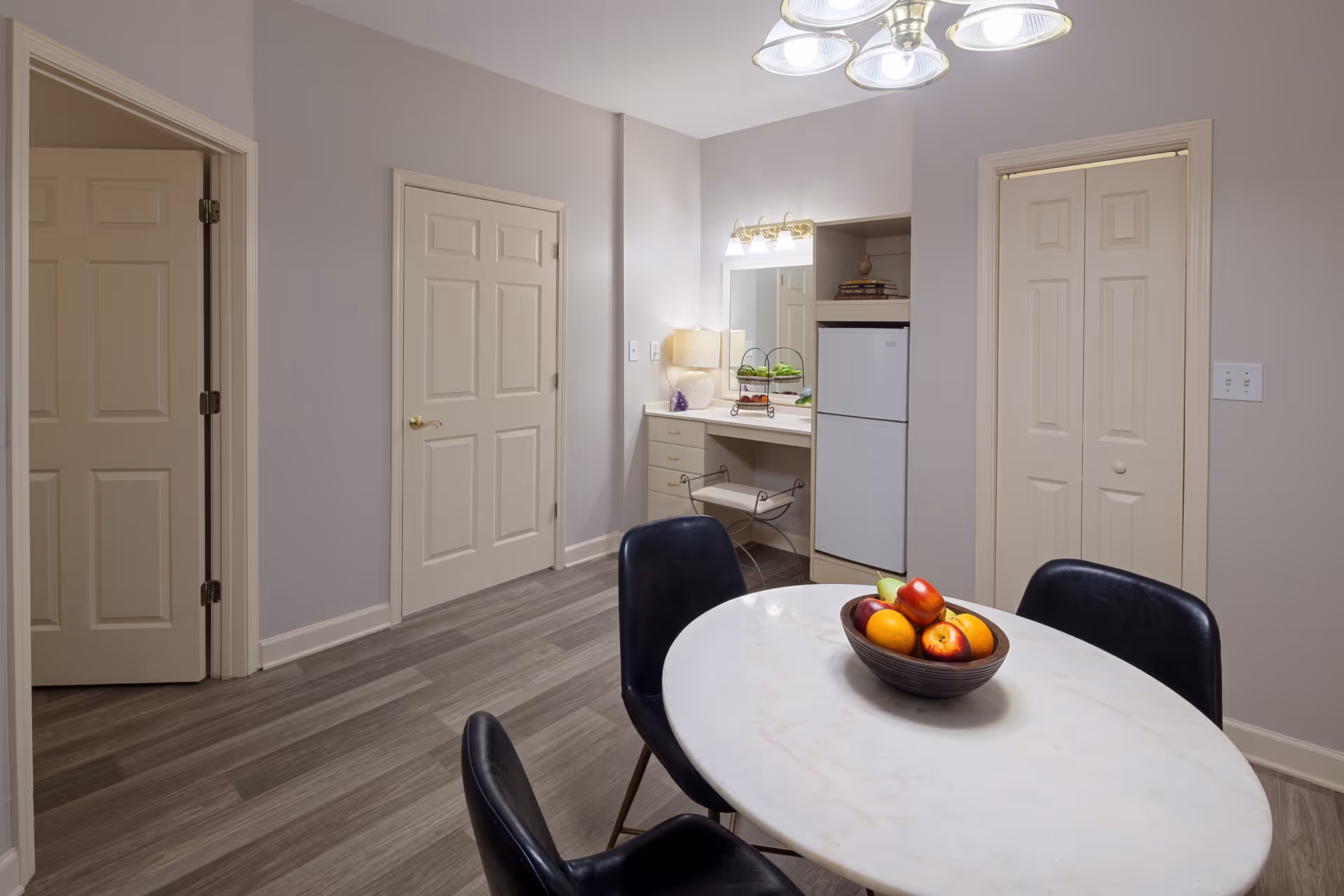 Interior view of a room with a round white table and four black chairs. On the table is a bowl filled with apples and oranges. The room has light gray walls and wood flooring. There are three closed doors, a small refrigerator, a vanity area with a mirror and light fixture, and a small lamp on the vanity.