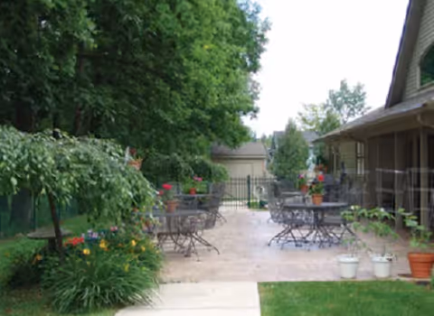 Outdoor patio area with several metal tables and chairs, potted plants on the tables and ground, surrounded by greenery and trees, adjacent to a building with a covered walkway.