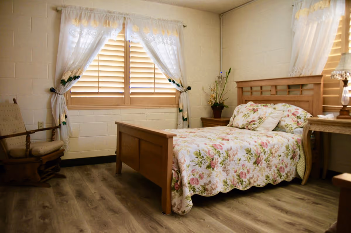 A cozy bedroom with a wooden bed frame and floral bedspread. The room has light-colored walls with two windows covered by wooden blinds and sheer white curtains with floral embroidery. There is a wooden rocking chair on the left and a wooden nightstand with a potted plant and a lamp on the right. The floor is covered with wood-like laminate.