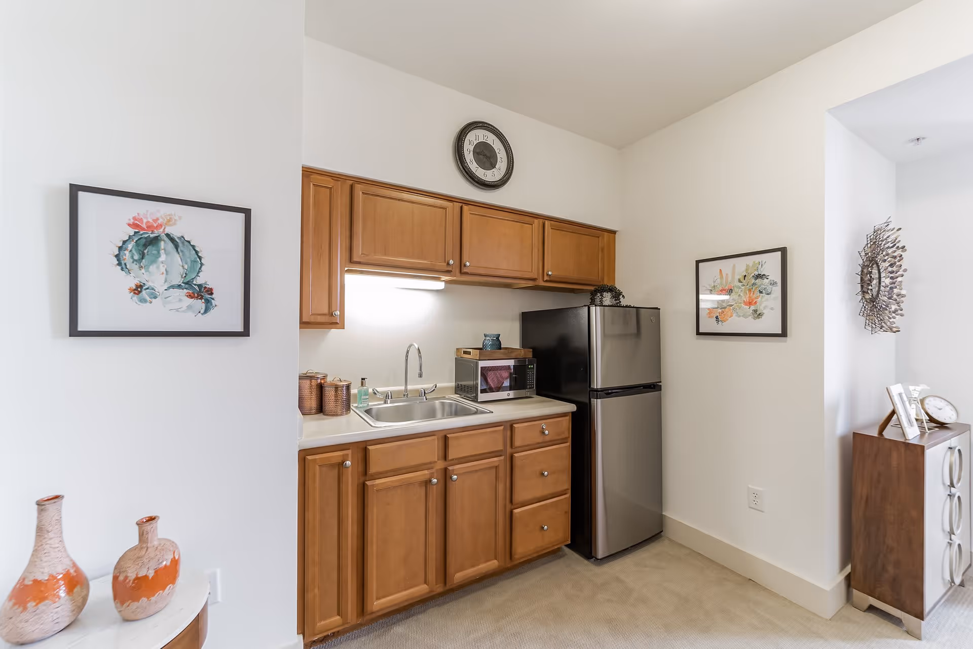 A small kitchen area with wooden cabinets, a stainless steel refrigerator, a microwave, and a sink. The walls are white and decorated with framed botanical artwork. There is a clock above the cabinets and decorative vases on a small table to the left.